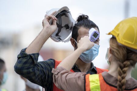 Factory woman worker in a face medical mask and safety dress used measures temperature at worker people standing on queue with a non-contact infrared thermometer.の写真素材
