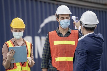 Factory man worker in a medical mask and safety cloth use equipment measures the worker temperature with a non-contact infrared thermometer.の写真素材