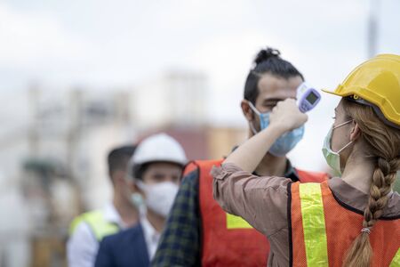 Factory woman worker in a face medical mask and safety dress used measures temperature at worker people standing on queue with a non-contact infrared thermometer.の写真素材