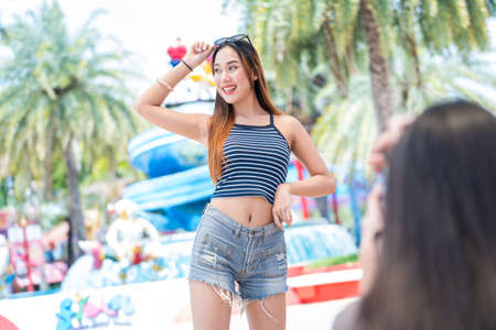 Young asian joyful woman in summer fashion dress standing and posing for friend take a shoot photo on smartphone in amusement park . Concept happy and lovely life of teenager.の写真素材