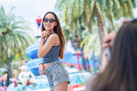 Young asian joyful woman in summer fashion dress standing and posing for friend take a shoot photo on smartphone in amusement park . Concept happy and lovely life of teenager.の写真素材