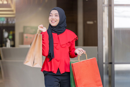 happy face muslim woman in red dress holding shopping bag and walking out shop building. Concept people activity in shopping festival season christmas and new year.の写真素材
