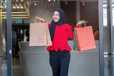 happy face muslim woman in red dress holding shopping bag and walking out shop building. Concept people activity in shopping festival season christmas and new year.の写真素材