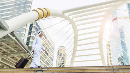 Business arab traveler saudi man carrying a suitcase and walk in the business landmark city with the modern steel structure and exterior wall modern buildingの写真素材