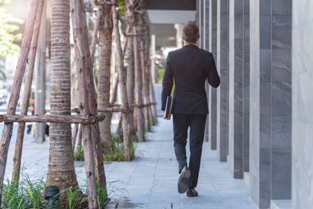 back of man in business suit walking and holding laptop notebook. business lifestyle in coffee time at morning daylight.の写真素材