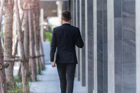 back of man in business suit walking and holding laptop notebook. business lifestyle in coffee time at morning daylight.の写真素材