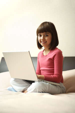 A caucasian girl sits on his bed, smiling and holding laptop. Beige background.の写真素材
