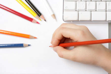 child draws on a white paper with colored pencils, a computer keyboard in the background