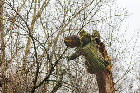 A moss-covered bear sculpture is perched on a tree branch, surrounded by trees in a cloudy environment.の写真素材
