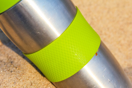 Stainless steel thermos bottle with bright green silicone band on sandy surfaceの写真素材