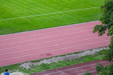 Red athletic running track surface with white lane markings alongside vibrant green grass sports...