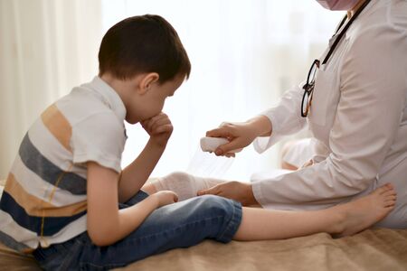 The doctor in a white coat carefully provides first aid. He puts a bandage on the boy's leg. The kid patiently, irritably, holds his chin with his hand and observes the process.の写真素材