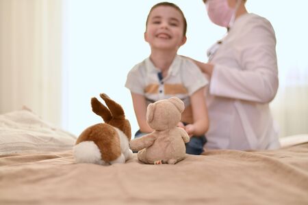 The child sits on the bed in front of toys and smiles contentedly. The doctor listens to the baby breath from the back.の写真素材