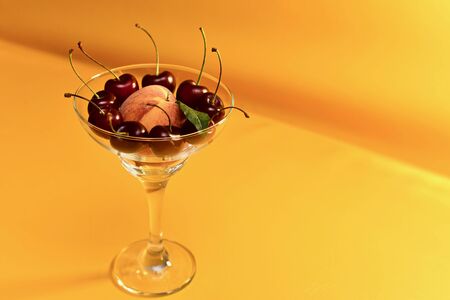 A vase of transparent glass with a thin stalk with fresh ripe peach and red cherries with stems. On a yellow background to the left. Top view from the front.の写真素材