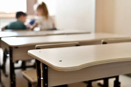 A row of school desks, the last empty tabletop in focus. At the first table sit a boy and a girl and chat. In the classroom, flooded with light from the window.の写真素材