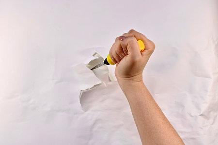 A screwdriver, with a yellow handle, in a tightly compressed woman's hand, stuck in a wall with a...