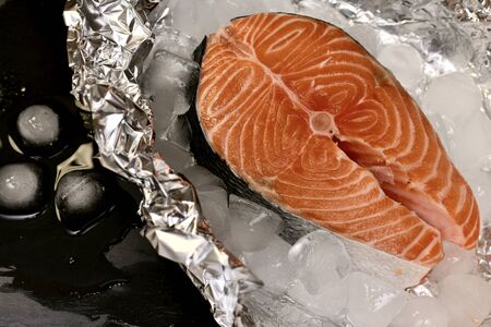 Red fish chilled with ice on foil next to thawed water on a black shale substrate.の写真素材