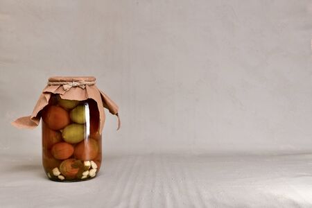 One glass jar covered with a lid and paper wrapper with a rope with canned fermented tomatoes, left on a gray cement background.の写真素材