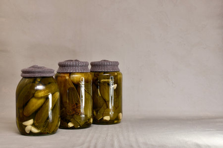 A diagonal row of three glass jars, with rust-proof knitted lids filled with fermented cucumbers,...