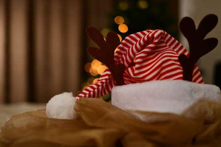 New Year red white striped cap with fur puffer and pompom, raised, with vertical deer horns, on festively draped fabric against the warm lights of the Christmas tree garland.の写真素材