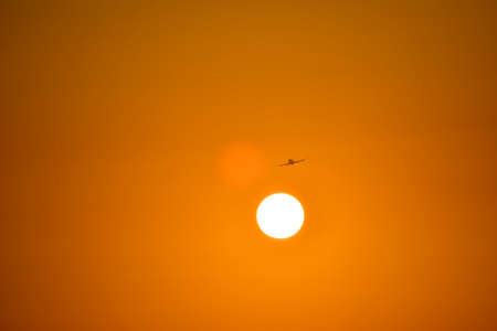 The silhouette of the plane on takeoff, with a fiery trace in the tail. It flies during the orange sunset. Flying around the flanked side, the white even circle of the sun. Towards the sunlight.の写真素材