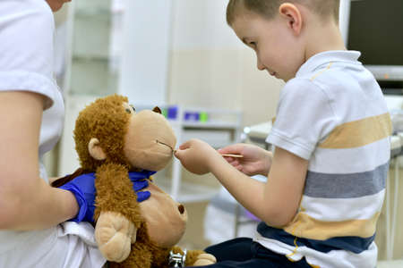 The child at the reception at the clinic examines the mouth cavity with a medical spatula of a toy plush monkey, which is in the hands of the doctor's blue protective gloves.の写真素材