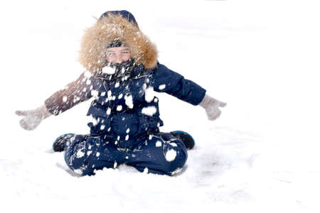 The child is warmly dressed, with a fur hood. Sitting on a frosty winter day, with his hands in mittens, he throws a lot many of collected snow up, scattering in the air.の写真素材