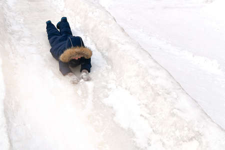 A child in winter clothes, in a fur hood and a hat with a pompom, cheerfully rolls down lying on his stomach, face forward, from a snowy mountain in winter.の写真素材