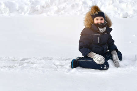 The child sits on the ice covered with snow, surrounded by snowdrifts. Dressed in winter clothes, slightly snowy places. On a clear sunny day.の写真素材