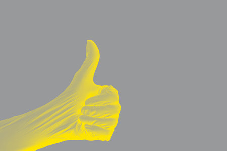 Gesture of a clenched fist of a hand with a thumb up extended upward in a protective stretched nitrile glove with yellow illumination on a uniform ultimate gray background. With gray tinted shadows.の写真素材