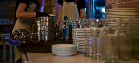 The working staff of the restaurant in uniform in aprons, sort stacks of clean shiny utensils in gloves in the background of the white plates, steel portion pans, glasses, tubes.の写真素材
