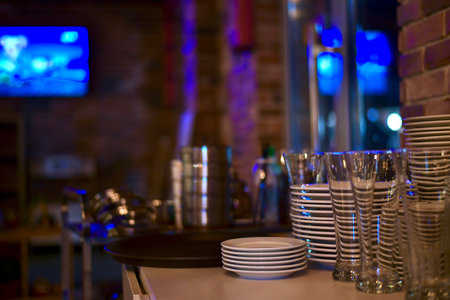 Stacks of a clean shiny utensils made of steel, ceramics and glass. On the countertop of the bollard near the brick wall, in a room with subdued lighting, and blue spots of light in the TV and window.の写真素材