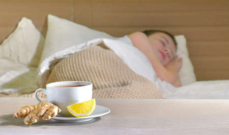 A cup of tea on a saucer with ginger root and a slice of lemon on a blurred background. Sleeping sick child in bed.の写真素材