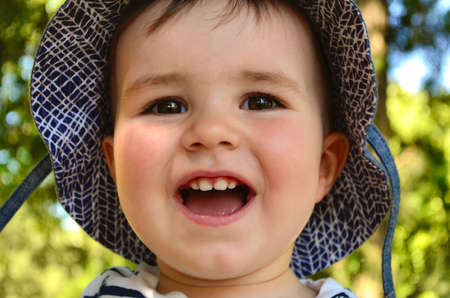 The happy child in the hat smiles. Emotion of joy and delight. Against the backdrop of summer greenery is a sunny day.の写真素材