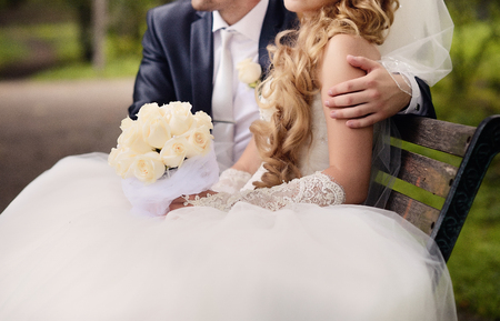 The bride and groom are sitting in the park on a bench. He embraces her, and in her hands a beautiful wedding bouquet of white roses.の写真素材