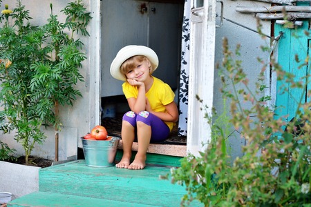 A small, pretty girl in a hat gathered a harvest of ripe red tomatoesの写真素材