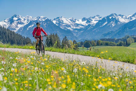pretty senior woman riding her electric mountain on warm spring day with snoe capped bike in the Iller valley near Oberstdorf with snoe capped Allgau High Alps in background, Allgau Alps, Bavaria,の写真素材