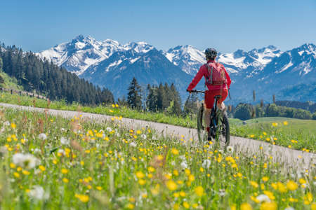 pretty senior woman riding her electric mountain on warm spring day with snoe capped bike in the Iller valley near Oberstdorf with snoe capped Allgau High Alps in background, Allgau Alps, Bavaria,の写真素材