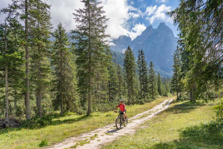pretty senior woman riding her electric mountainbike in the Fischlein Valley in the Sexten Dolomites wit famous Sextan sundial in background, Tre cime National park, South Tirol, Italyの写真素材