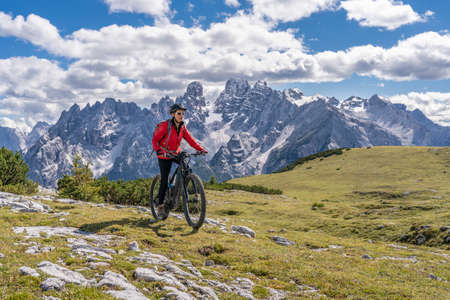 nice and active senior woman riding her electric mountain bike on the high plateau of Pratto Piazzo in the three peaks Dolomites, rocky silhouette of Mount Cristallo in background, South Tirol, Italyの写真素材