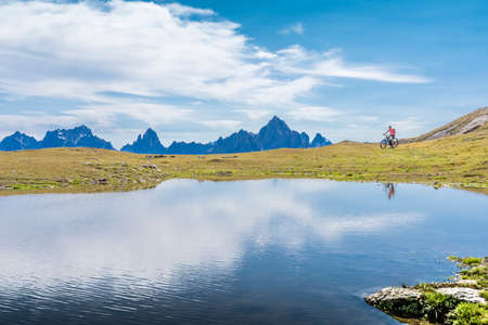 nice woman riding her electric mountain bike the Three Peaks Dolomites, reflecting herself in the blue water of a cold mountain lakeの写真素材