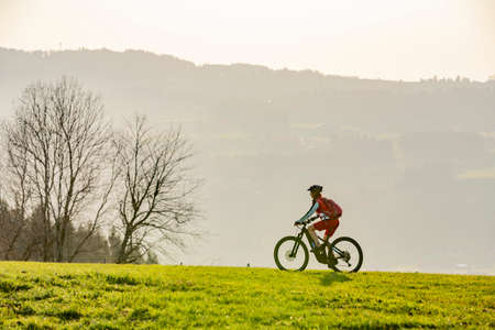 pretty senior woman riding her electric mountain bike in early springtime in the Allgau mountains near Oberstaufen, in warm evening light below the spectacular snow capped mountains of Nagelfluh chainの写真素材