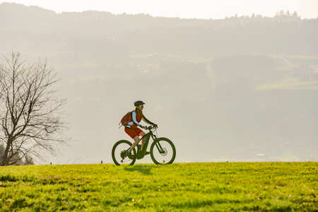 pretty senior woman riding her electric mountain bike in early springtime in the Allgau mountains near Oberstaufen, in warm evening light below the spectacular snow capped mountains of Nagelfluh chainの写真素材