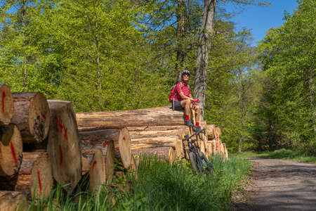 nice and remained young senior woman, sitting and having a rest break on a pile of tree trunks during a bicycle tour in a springtime forest in the Stromberg area near Zaberfeld, Baden-Wuerttemberg,の写真素材
