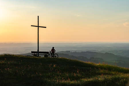 pretty senior woman riding her electric mountain bike in warm dawn sunlight below the summit cross of Salmas hight above Oberstaufen, Allgau Alps, Bavaria Germanyの写真素材