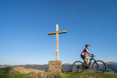 pretty senior woman riding her electric mountain bike in warm dawn sunlight below the summit cross of Salmas hight above Oberstaufen, Allgau Alps, Bavaria Germanyの写真素材