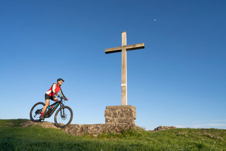 pretty senior woman riding her electric mountain bike in warm dawn sunlight below the summit cross of Salmas hight above Oberstaufen, Allgau Alps, Bavaria Germanyの写真素材