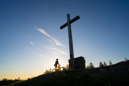 pretty senior woman riding her electric mountain bike in warm dawn sunlight below the summit cross of Salmas hight above Oberstaufen, Allgau Alps, Bavaria Germanyの写真素材