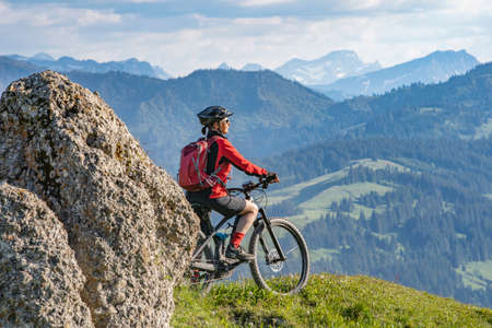 pretty senior woman riding her electric mountain bike in warm dawn sunlight and enjoying the spectacular view over the Allgau alps near Oberstdorf, Bavaria, Germanyの写真素材