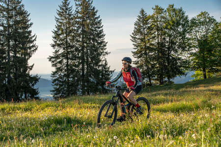 pretty senior woman riding her electric mountain bike on the mountains above Oberstaufen, Allgau Alps, Bavaria Germany, view into the Bregenzer Wald mountain range in Vorarlberg, Austriaの写真素材